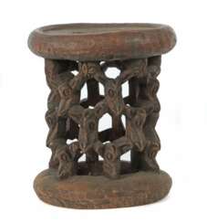 Afrikanischer Hocker aus einem Stück Holz geschnitzt, mit ringförmigem Stand und gitterartig angeordneten Miniaturmasken als Stützen der leicht ovalen Sitzfläche