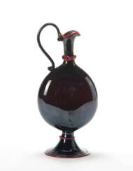 (Attributed) | Small dark amethyst blown glass pourer, iridescent on the outer surface and decorated with red glass paste applications. Murano, 1920s. (h 25 cm.)