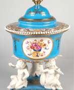 Produits en porcelaine. Prunkdeckelgefäß, wohl Frankreich um 1900, dreipassiger Fuß mit drei Putten mit Muschelhorn auf Delphinen sitzend, Aufsatz mit goldgerahmten Blumenmedaillons, Deckel und Wandung mit reichem Golddekor, Bodenmarke in der…