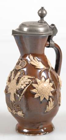 Kleine Kanne mit Zinndeckel, datiert 1777, braun glasiert mit hellen floralen Auflagen, Schauseite mit bekröntem Doppeladler mit Zepter und Reichsapfel, darunter Monogr. I.A.R.H., Kugeldaumendrücker, Ausguß und Stand bes… - photo 1