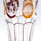 Becherglas, Böhmen, untere Hälfte facettiert geschliffen, im oberen Teil 6 Medaillons mit eingeschliffenen floralen und figürlichen Motiven, z.T. bernsteinfarben und dunkelrot lasiert, Goldrand, H. 13,5 cm - photo 1