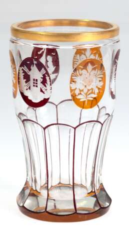 Becherglas, Böhmen, untere Hälfte facettiert geschliffen, im oberen Teil 6 Medaillons mit eingeschliffenen floralen und figürlichen Motiven, z.T. bernsteinfarben und dunkelrot lasiert, Goldrand, H. 13,5 cm - photo 1