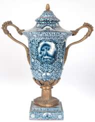 Deckel-Vase, Keramik mit Bronzebesatz, vollflächiger Blaudekor, auf quadratischem Sockel reliefierter Bronze-Fuß, seitl. Bronze-Henkel mit Beeren- und Akanthusblattdekor, Bronze-Pinienzapfen als Deckelbekrönung. H. 54 cm…