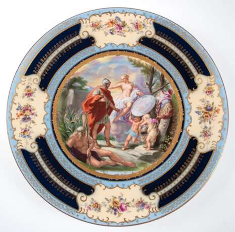 Großer Wandzierteller, Porzellanfabrik Eichwald, Böhmen um 1900, im Spiegel mythologische Szene, lithographischer Vordruck, polychrom staffiert, Fahne mit blauen Bändern mit goldenem Ornamentdekor und 4 Kartuschen mit Bl… - Foto 1