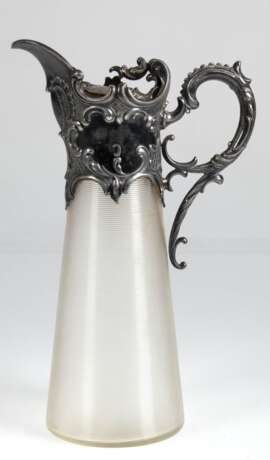 Historismus-Schenkkanne mit versilberter Metallmontierung, um 1880, konischer Glaskorpus mit horizontalem Rillendekor, reich reliefierte Metallmontur mit Scharnierdeckel und hochgezogenem Ohrenhenkel, H. 29,5 cm - photo 1 Historismus-Schenkkanne mit versilberter Metallmontierung, um 1880, konischer Glaskorpus mit horizontalem Rillendekor, reich reliefierte Metallmontur mit Scharnierdeckel und hochgezogenem Ohrenhenkel, H. 29,5 cm - photo 1